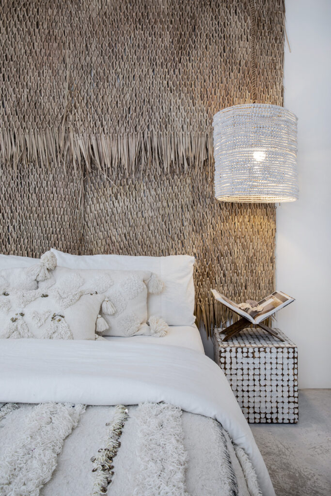 Boho-style bedroom with a textured woven headboard, white bedding with tassel pillows, a beaded nightstand, and a woven pendant light creating a cozy, natural aesthetic.