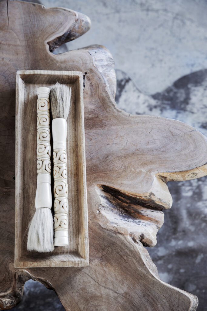 Rustic wooden table with a carved wooden tray holding two decorative calligraphy brushes with intricate ivory-like handles and soft bristles.