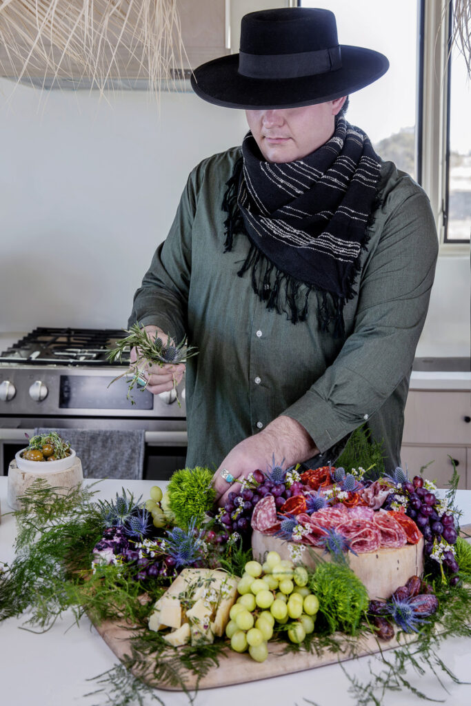 Man in a black hat and scarf styling a gourmet charcuterie board with grapes, cheese, and cured meats in a rustic kitchen setting.