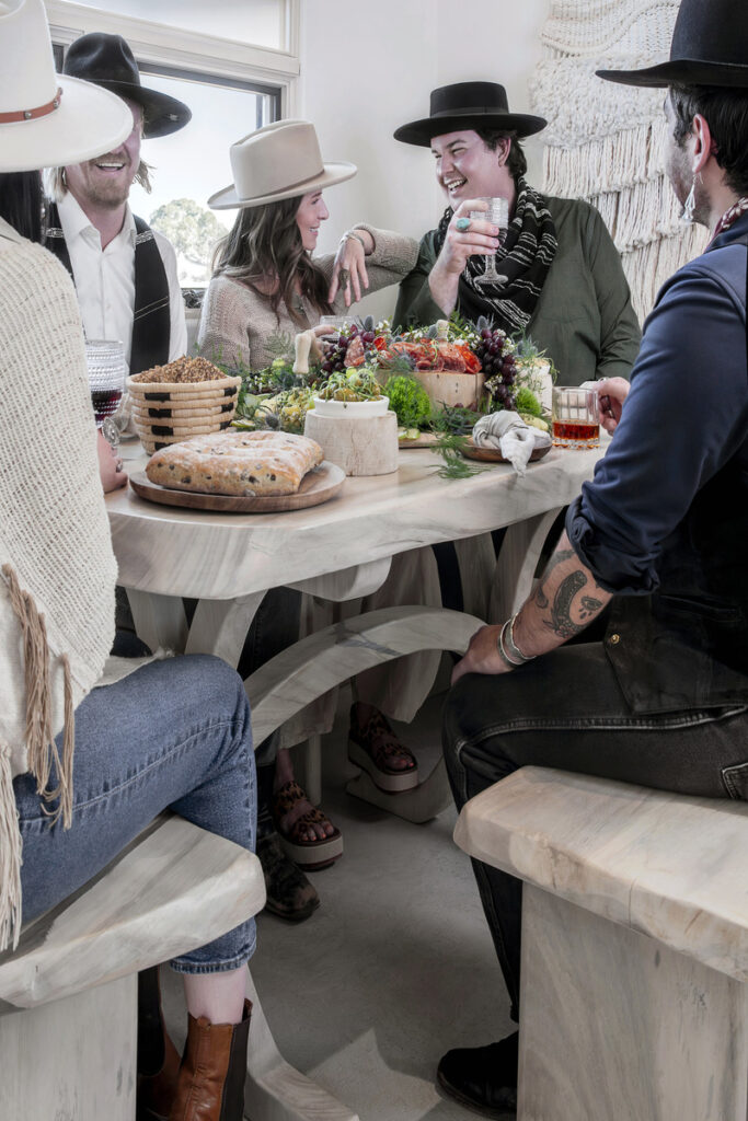 Group of stylish friends in cowboy hats enjoying a rustic charcuterie feast at a wooden dining table, sharing drinks and laughter.