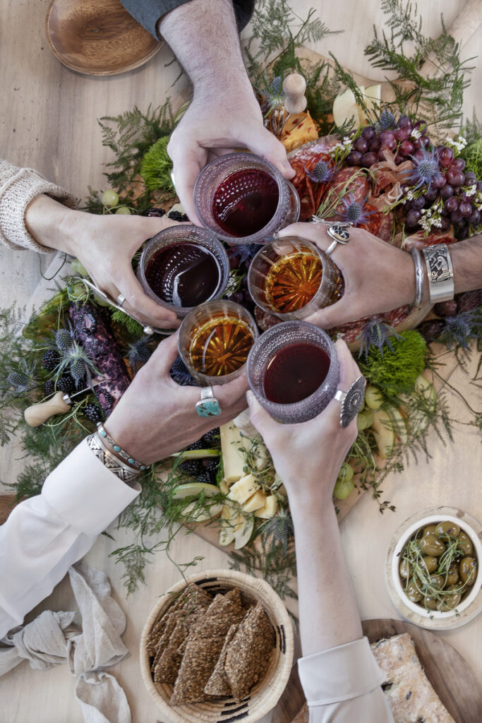 Close-up of friends toasting with drinks over a beautifully arranged charcuterie board.