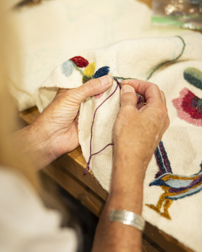 A Stitch in Time Close-up of hands embroidering a traditional colcha stitch on wool fabric, featuring colorful floral and bird motifs, representing the rich heritage of New Mexico textile art.