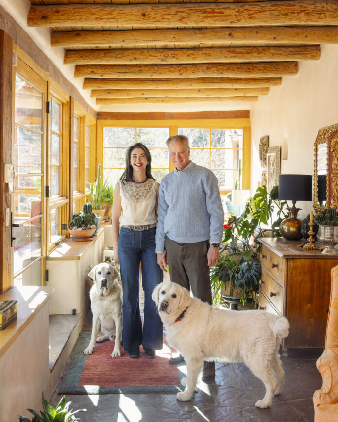 Adelma and Tom Hnasko stand in their historic Santa Fe adobe home on Upper Canyon Road, surrounded by Southwestern decor, natural light, and their two large dogs.