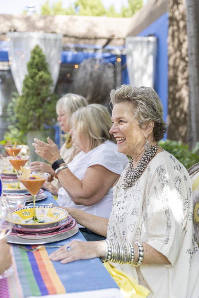 A group of women enjoying a lively outdoor lunch gathering, seated at a colorful table adorned with plates of food and drinks.