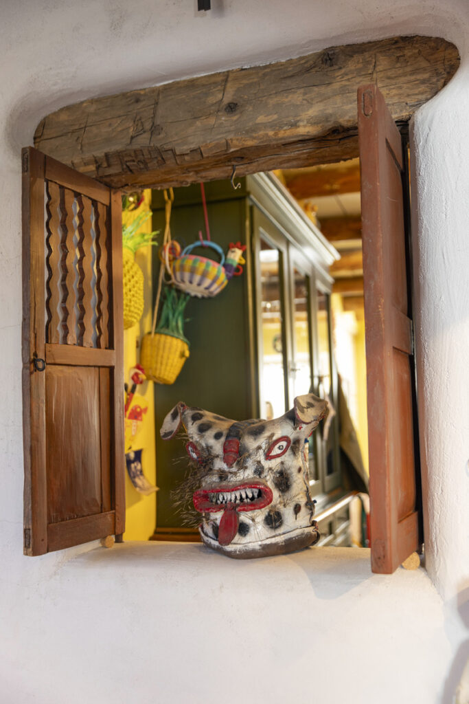 Rustic adobe home interior with a traditional wooden window, showcasing a hand-carved tribal mask on the windowsill and colorful woven baskets in the background.