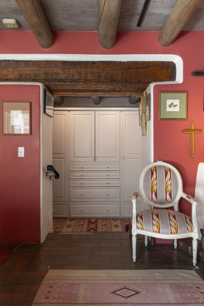 Southwestern-style home interior with exposed wooden ceiling beams, red adobe walls, and a vintage white armchair with patterned upholstery.