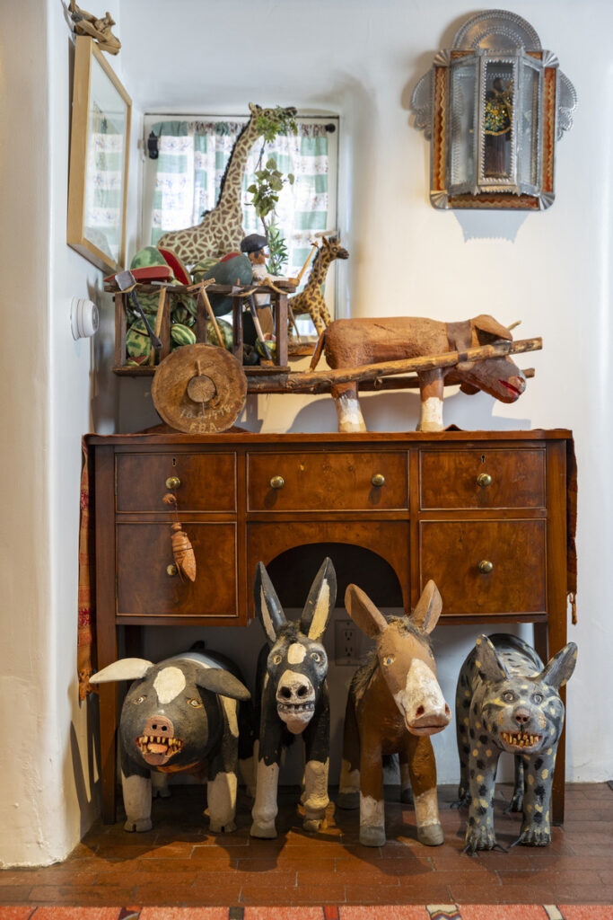 Rustic wooden animal sculptures, including donkeys, a pig, and a spotted dog, displayed under a vintage wooden cabinet with a handcrafted ox cart and giraffe figurines in a traditional Mexican folk art setting.