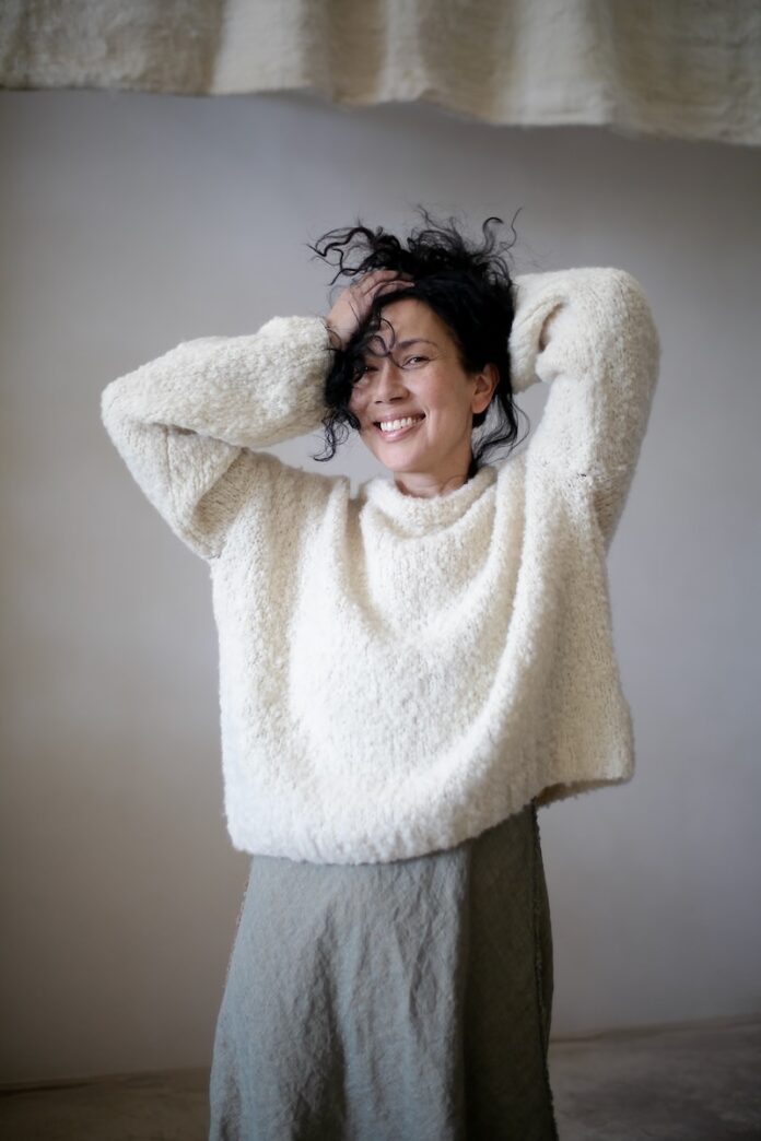 A woman with black hair in a white sweater and grey pants poses, representing Santa Fe style through fashion.
