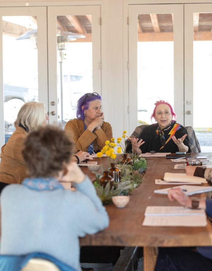 A trip down memory lane where people gathered around a rustic wooden table in a bright, modern space, engaged in a discussion. A woman with pink hair speaks expressively while others listen attentively, taking notes.