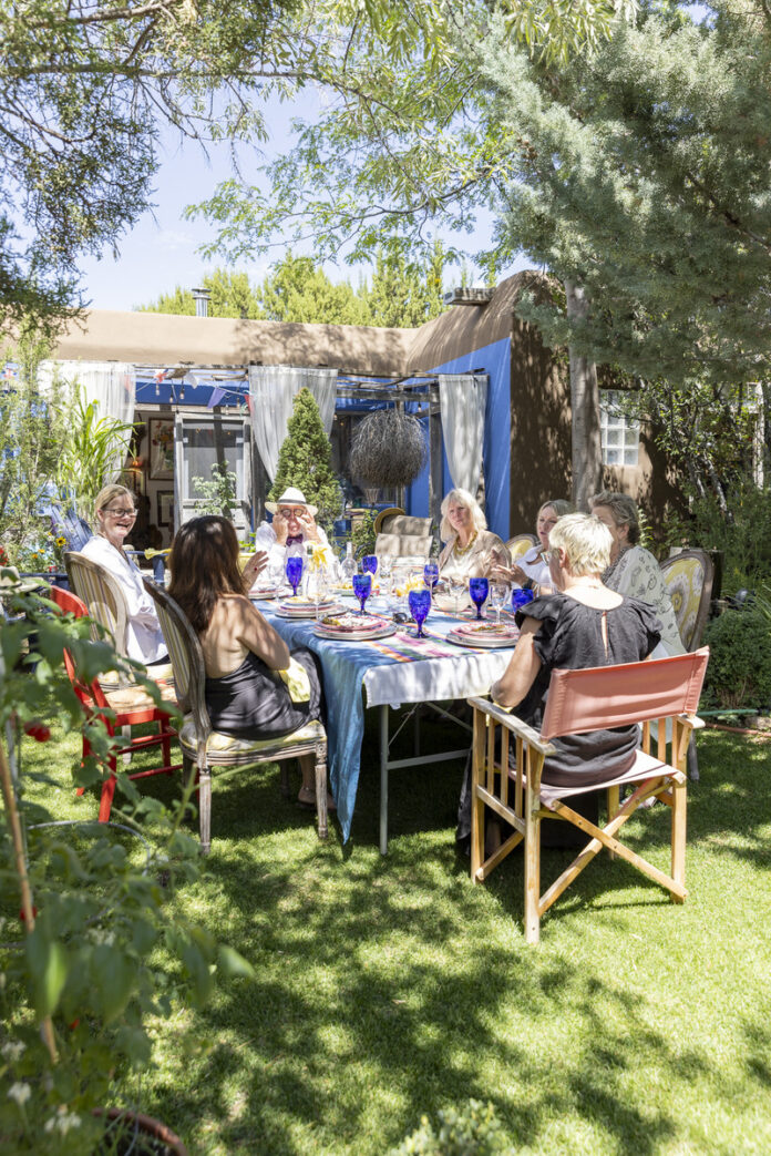 A group of women enjoying an outdoor lunch in a garden setting, seated around a table adorned with colorful glasses and plates, amidst lush greenery and a relaxed atmosphere.