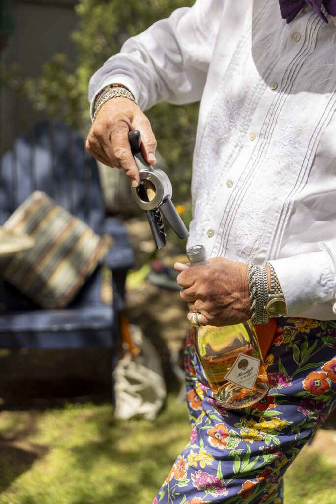 A person in a white shirt and colorful floral pants using a corkscrew to open a bottle in a relaxed outdoor setting.