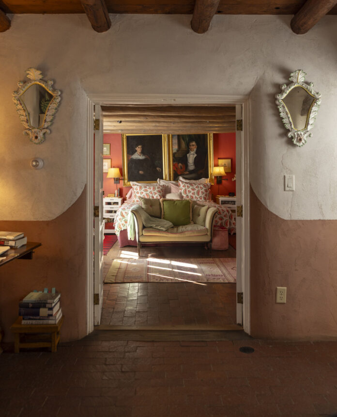 Rustic Santa Fe-style bedroom with wooden ceiling beams, adobe walls, and vintage decor, featuring a cozy seating area and antique portraits.