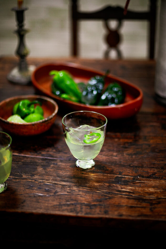 Santa Fe-style cocktail with mezcal, absinthe, and poblano garnish on a rustic wooden table, surrounded by fresh limes and peppers.