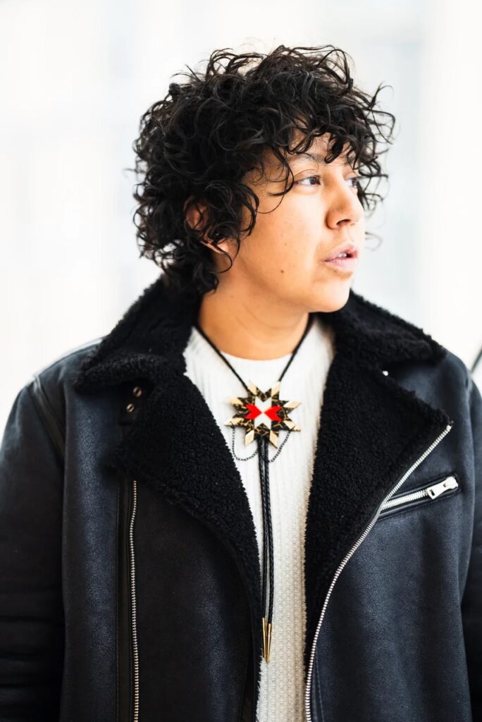 A person in a black leather jacket with black ruffled hair and a bollo tie over their white shirt.
