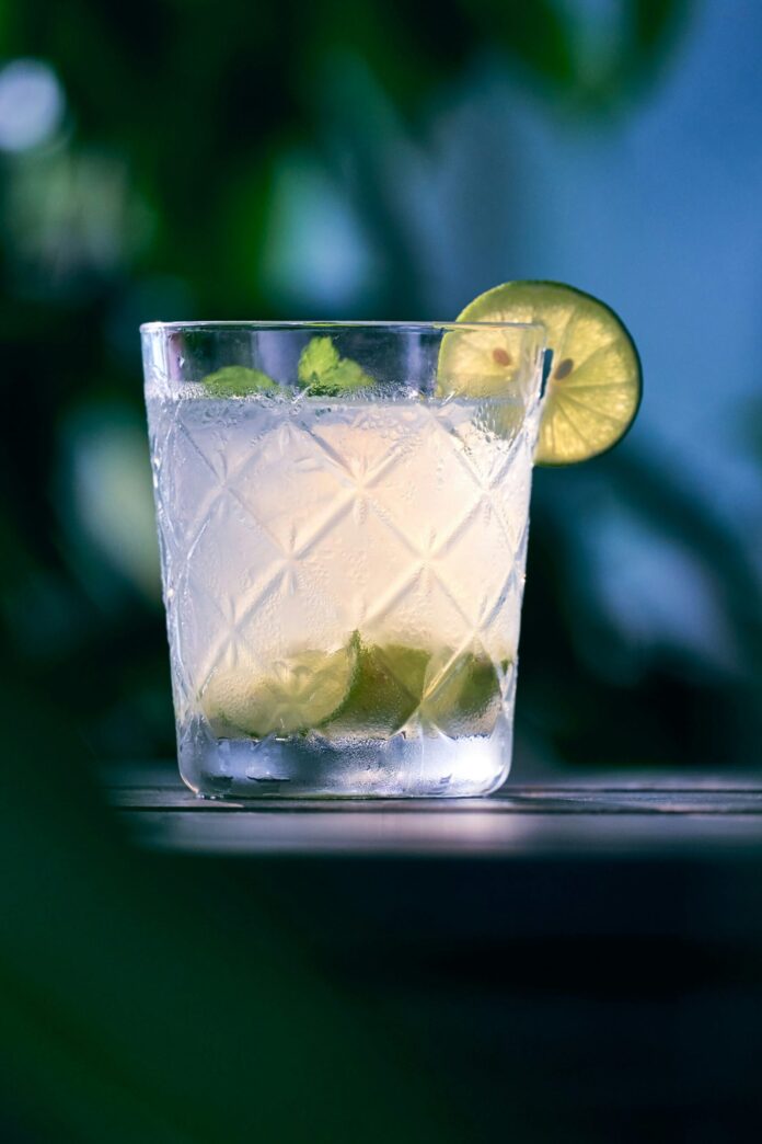 A glass of tequila blanco sits on a table with a wedge of lime inside and a garnish of lime and mint.