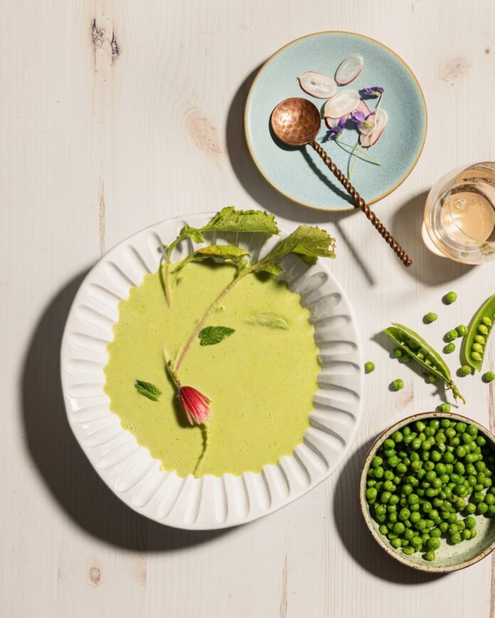 A white bowl holds a Chilled Pea Soup with radish and mint garnishes as a bowl of peas sits off to the right.