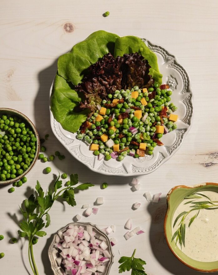 A white bowl holds butter lettuce leaves with plenty of peas, cheddar cheese, and bacon. Off to the bottom right corner sits a bowl of green goddess dressing.