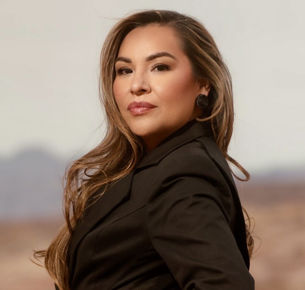 A woman with long brown hair stands in the desert in a black suit jacket.
