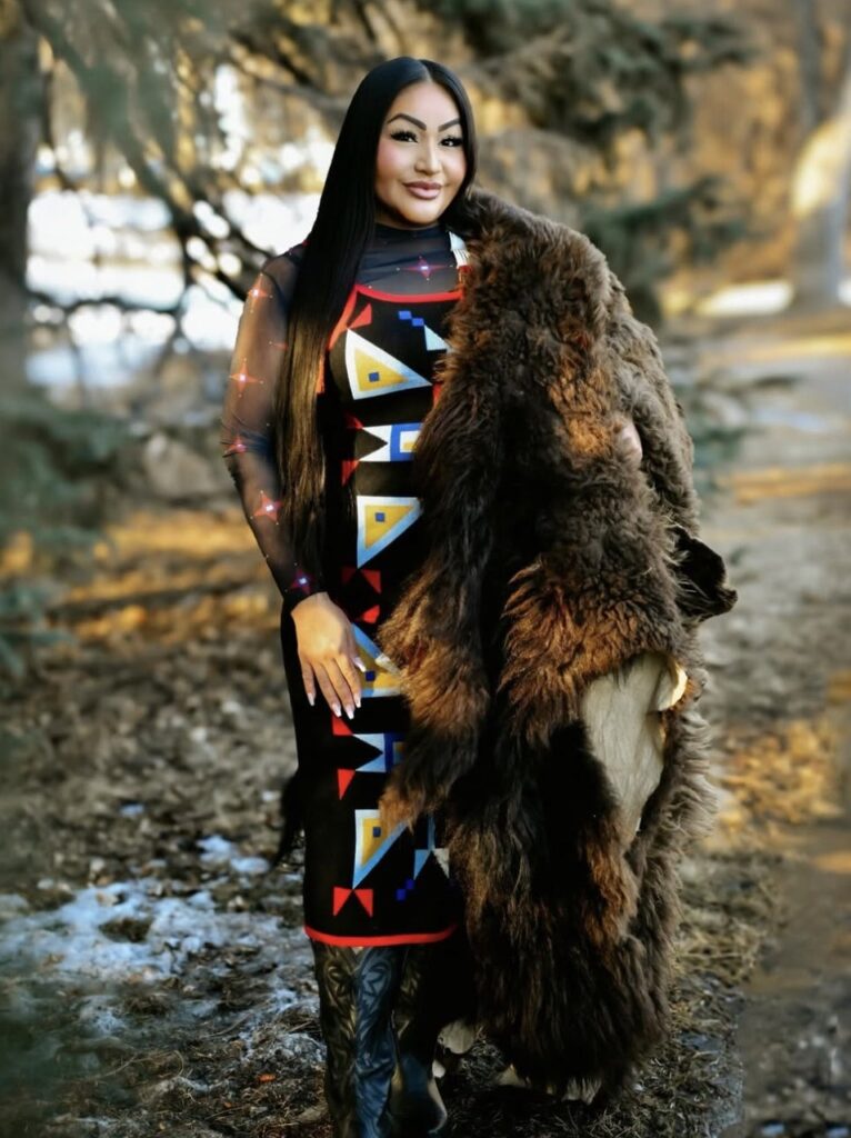 A woman in a native dress holds a bear pelt over her shoulder in the woods.