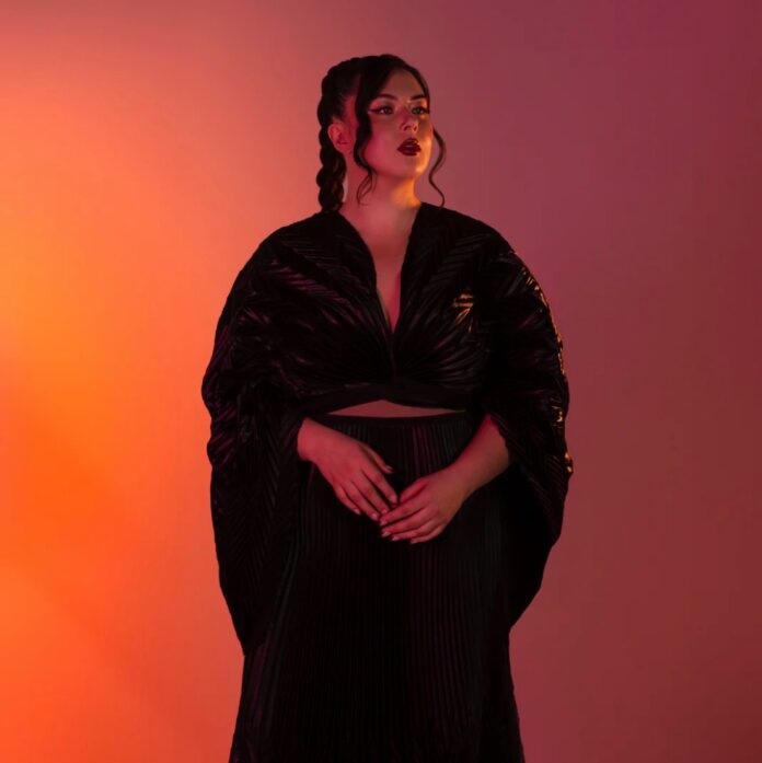 A woman in a native black dress stands in front of an orange and maroon backdrop with her hands folded in front of her torso.