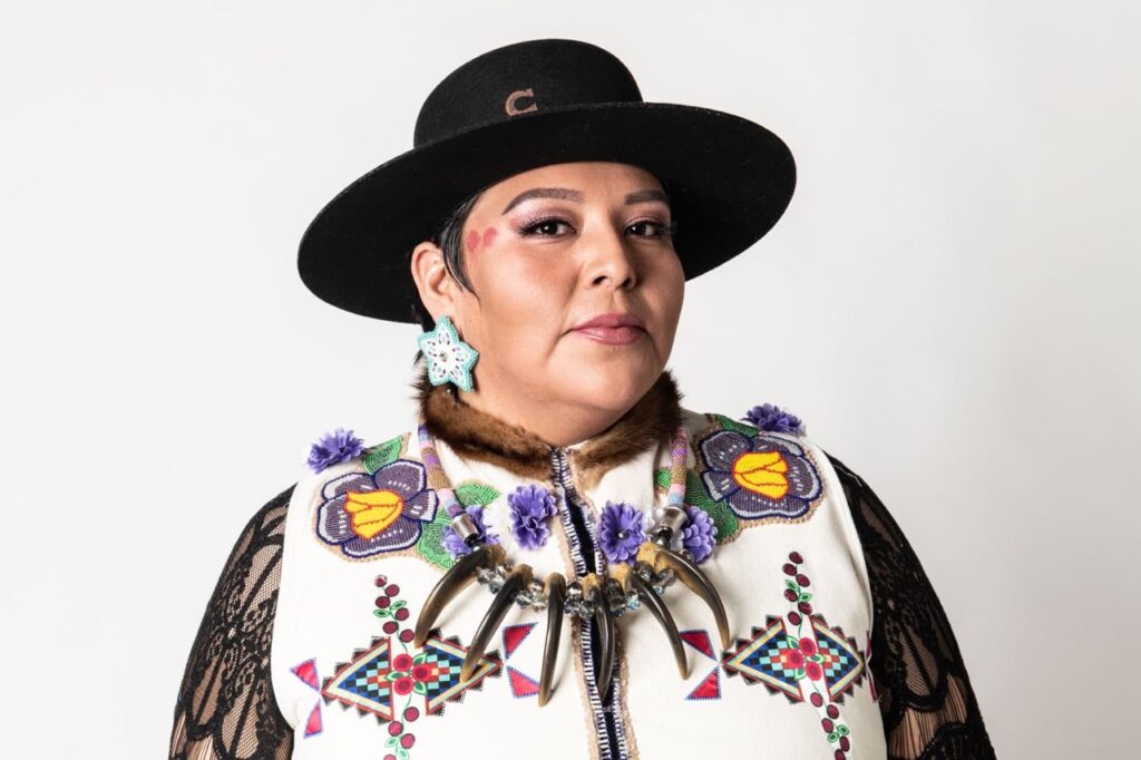 A person in native dress and a bone necklace stands with large flower earrings and a black hat on as well.