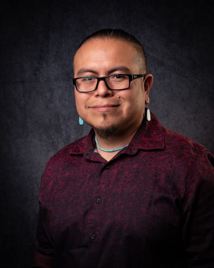 A man in a maroon polo shirt sits in front of a grey background in black glasses and gold earrings.
