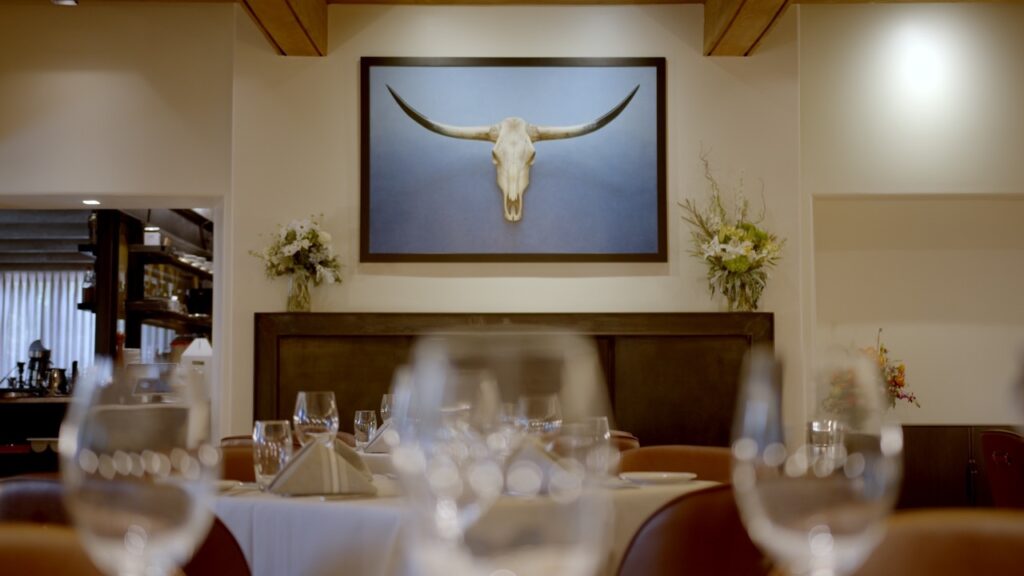 A painting of a longhorn skull sits on the wall at Market Steer Steakhouse with wine glasses on the table below it.