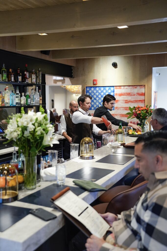 Two male bartenders mix up cocktails at Market Steer Steakhouses' bar as two male customers sit on the other side.