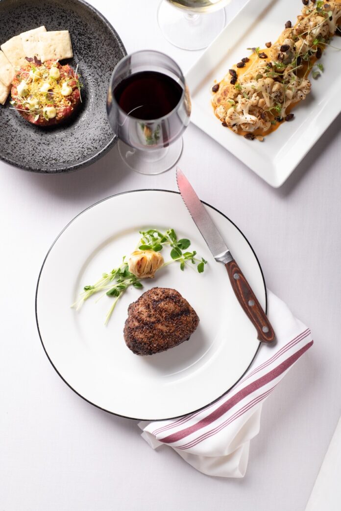 A white plate holds a piece of steak from Market Steer Steakhouse with a dark plate side and white rectangular plate appetizer in the top corners.