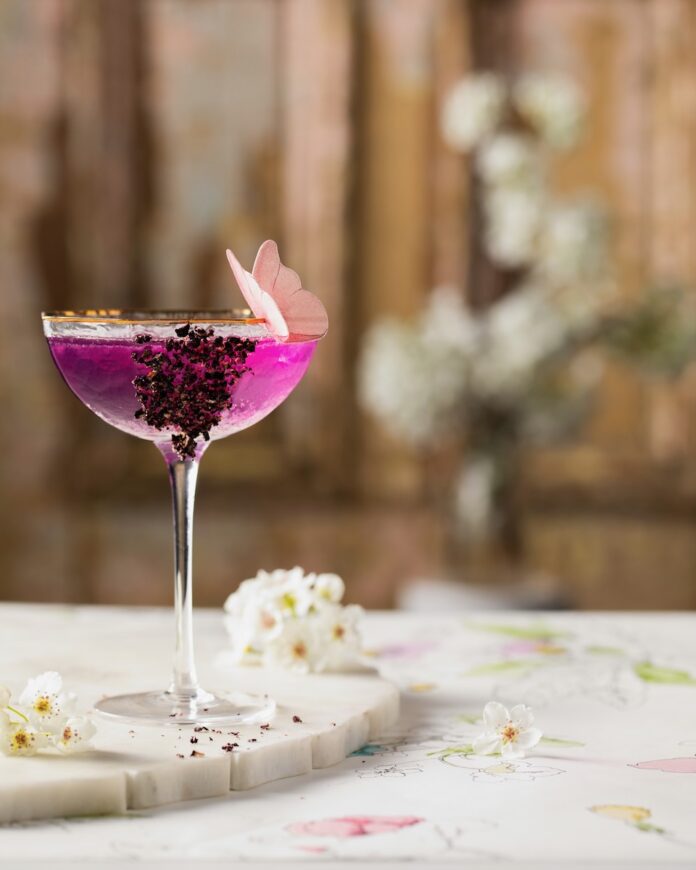 A coupe glass with a purple Mother's Day Gin and Tonic mocktail with crushed freeze-dried blueberries on the side and an edible pink butterfly on the rim. White flowers are scattered nearby.