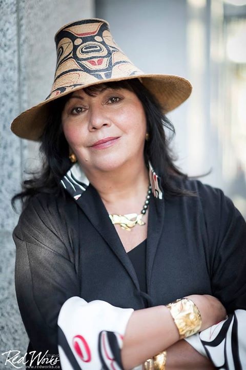 A woman with a tall hat and earrings stands with her arms crossed in a black shirt.