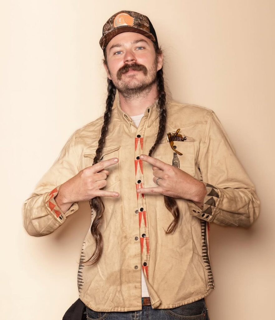 A man in a brown button up shirt shows off horns on his hands with long braids, a mustache, and a trucker hat on.