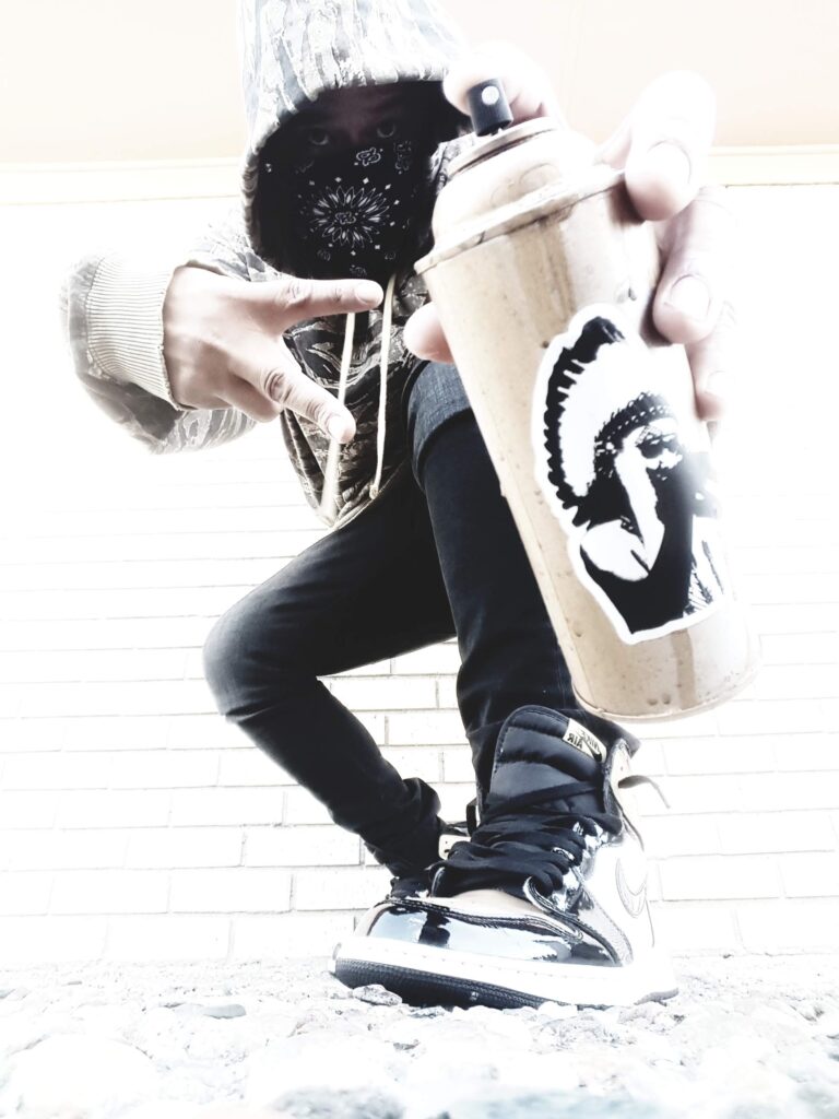 A man bends down in skinny jeans and a hoodie, holding a spray paint can up to the camera.