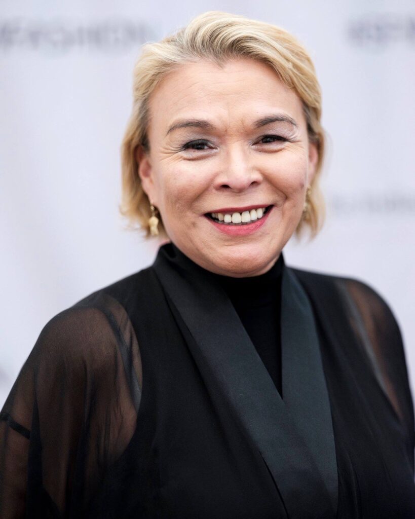 A woman in a black blouse and gold earrings stands in front of a white wall.