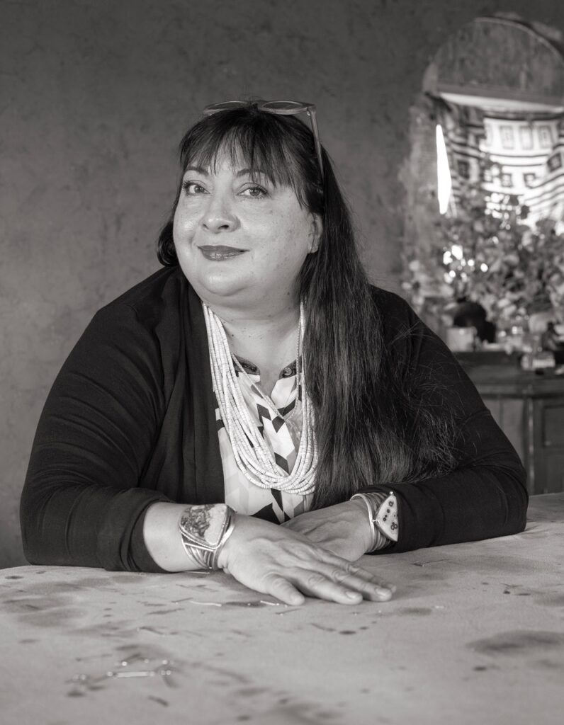 A woman in black and white sits at a wood table in a black jacket.