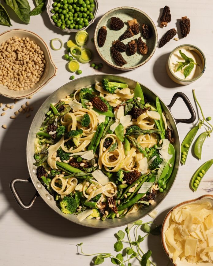 In a large steel pot sits a Pea Pasta Primavera with peas and other ingredients surrounding the pot on a white table.