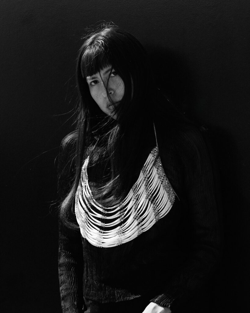 A woman in black native wears stands in the shadows with her hair covering half her face.