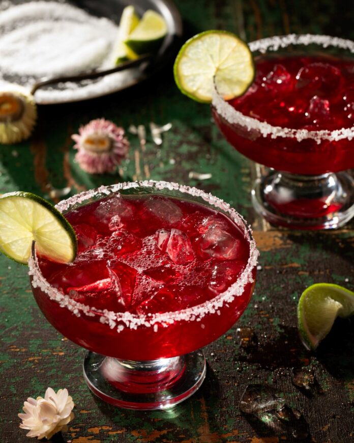 Two margarita glasses with salt rims are filled with a red pomegranate margarita with a lime wheel garnishing each glass.