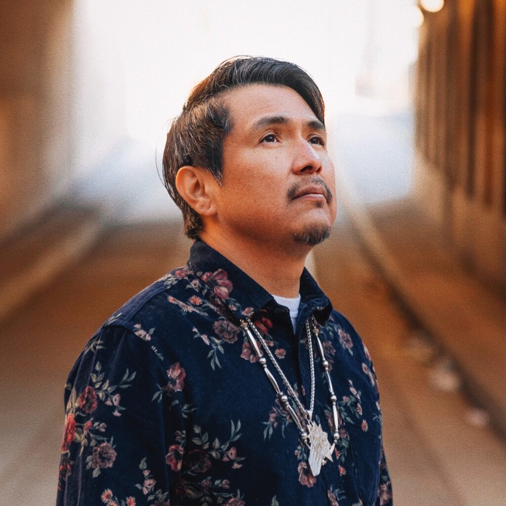 A man in a button up blue shirt with a native style necklace looks up to the sky.