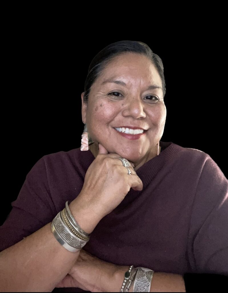 A woman in a maroon shirt smiles with her hand under her chin.