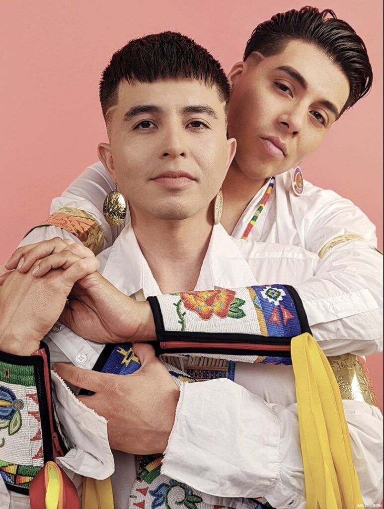 One man wraps his arms around the other from behind while both wear native styled white shirts.