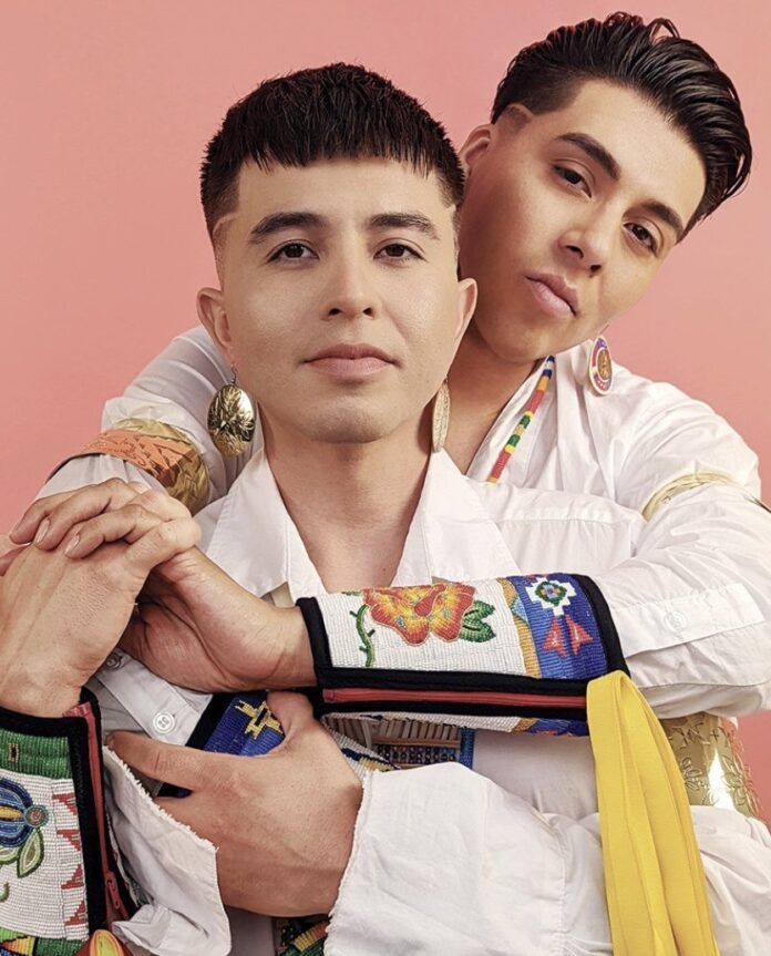 One man wraps his arms around the other from behind while both wear native styled white shirts.