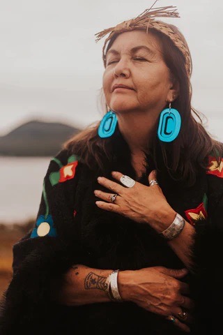 A woman in a sweater and blue earrings stands outside with her hand over her heart.