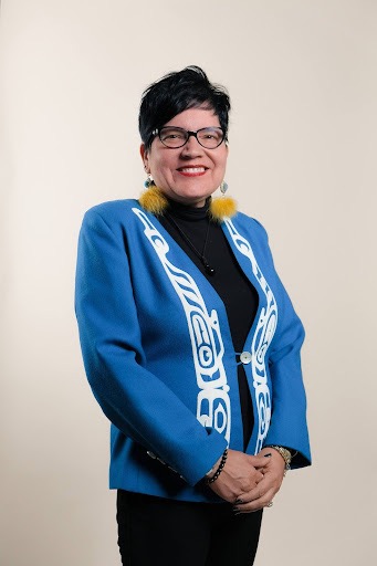 A woman in a native blue shirt jacket and big yellow earrings stands in front of a white background.