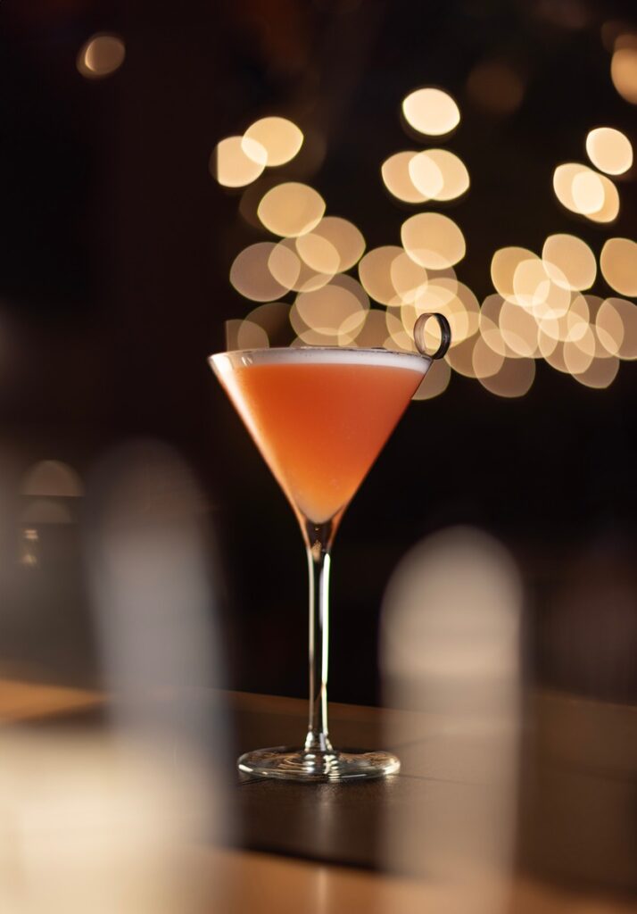 A tall martini glass with a vibrant orange cocktail sits on a polished bar surface.
