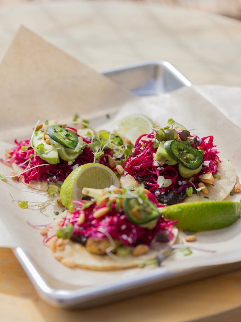 Three brightly colored tacos with shredded red cabbage, green toppings, and lime wedges are presented on a metal tray lined with parchment paper.