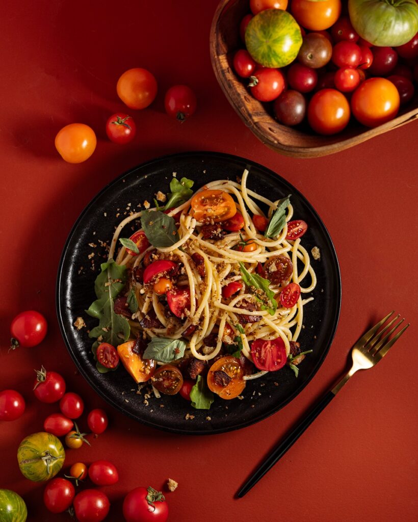 A bowl of BLT pasta with golden spaghetti, bacon pieces, red and yellow halved cherry tomatoes, and fresh green basil leaves.