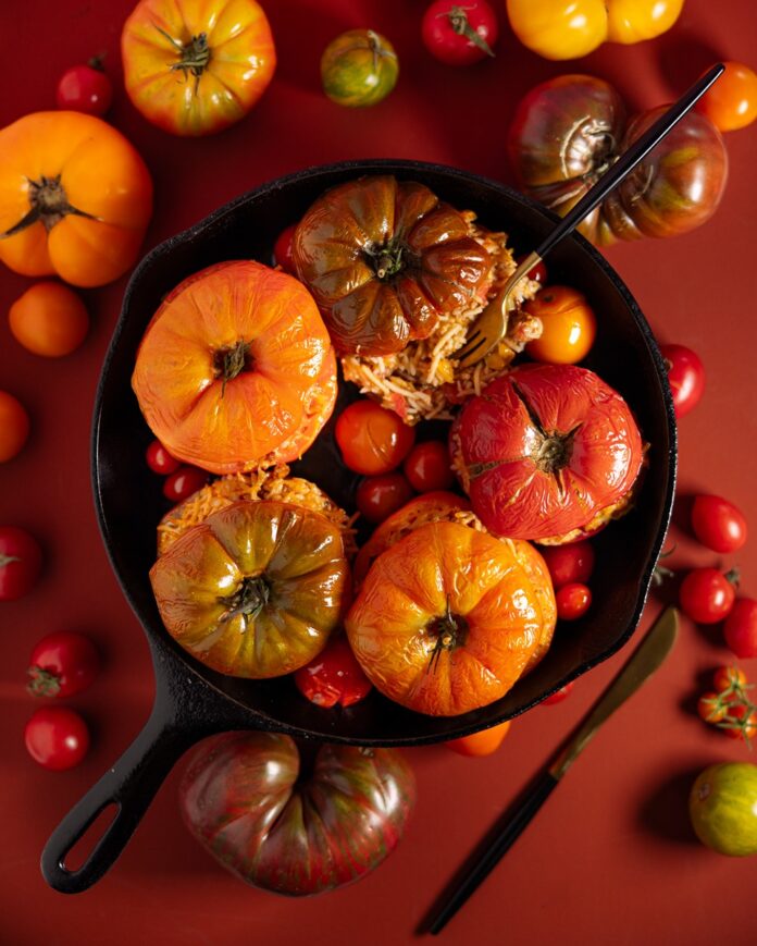Baked tomatoes, some with their tops removed and others still lidded, are arranged in a dark cast-iron skillet, surrounded by smaller, colorful cherry tomatoes and green herbs.