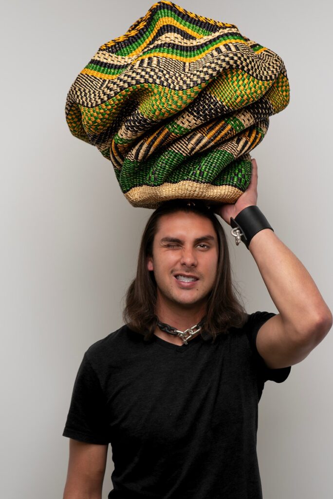 A person with long brown hair winking while balancing a large, colorful woven Bolga basket on his head.