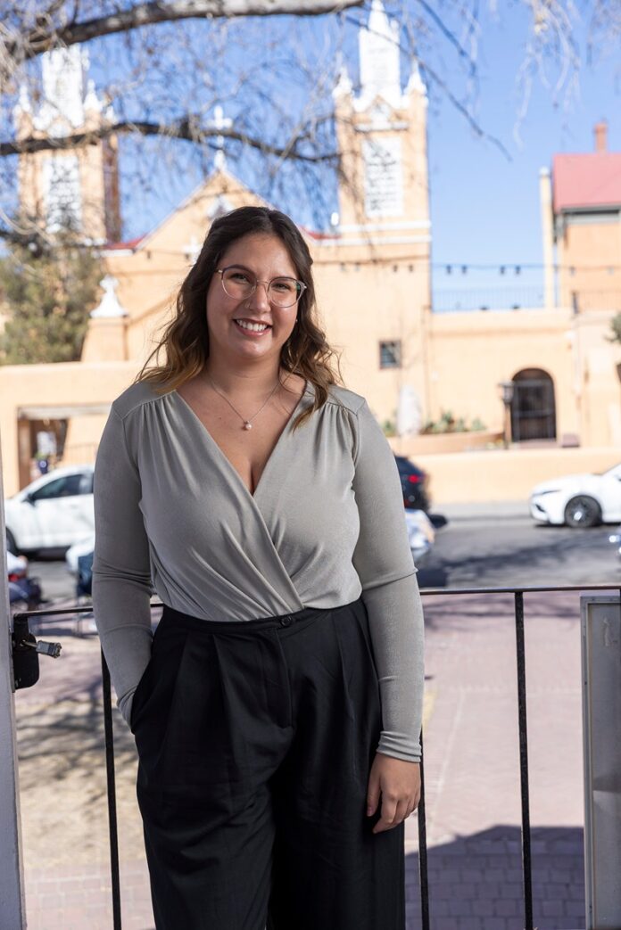 Brissa Chilton-Garcia, stands in traditional Spanish market on a balcony with a light-colored building featuring church-like spires and arches in the background